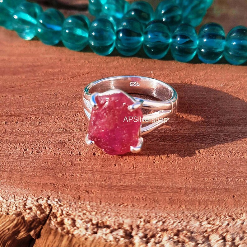 K&ouml;nnte beinhalten: Ein silberner Ring mit einem gro&szlig;en, rohen Rubin-Edelstein. Der Ring hat eine geteilte Schiene und ist mit "S26" und "APS Silvershop" gekennzeichnet.