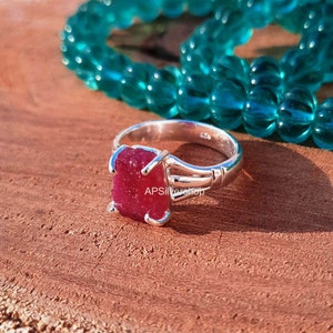 K&ouml;nnte beinhalten: Ein silberner Ring mit einem gro&szlig;en, rohen, roten Edelstein. Der Ring liegt auf einer Holzoberfl&auml;che mit einer t&uuml;rkisfarbenen Perlenkette im Hintergrund.