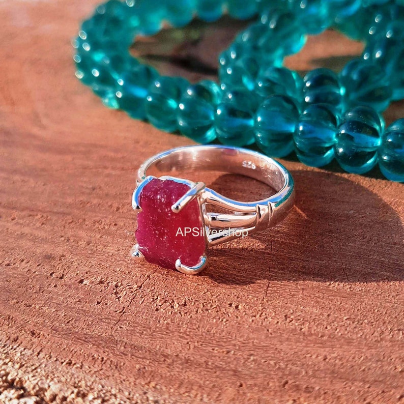 K&ouml;nnte beinhalten: Ein silberner Ring mit einem gro&szlig;en, rohen, roten Edelstein. Der Ring liegt auf einer Holzoberfl&auml;che mit einer t&uuml;rkisfarbenen Perlenkette im Hintergrund.