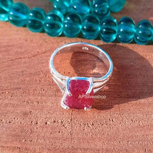 K&ouml;nnte beinhalten: Ein silberner Ring mit einem roten Edelstein, der in einer Krappenfassung sitzt. Der Ring liegt auf einer Holzoberfl&auml;che. Der Text "APSilvershop" ist auf dem Ring sichtbar.