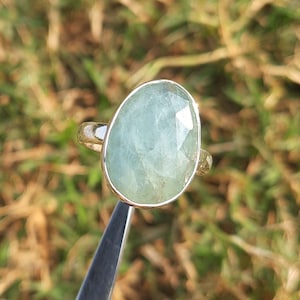 May include: A silver ring featuring a large, oval-shaped aquamarine gemstone. The gemstone is a pale blue-green colour and has a faceted cut.