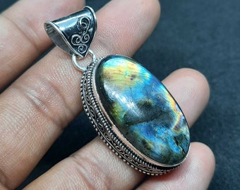Puede incluir: Pendiente de plata con una piedra preciosa de labradorita de forma ovalada. La piedra preciosa presenta un brillo iridiscente azul, verde y dorado. El pendiente está adornado con un diseño de plata intrincado alrededor del borde.