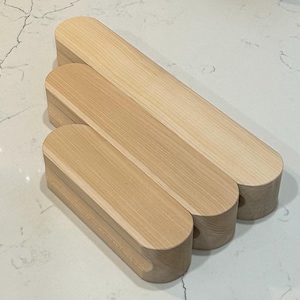 May include: Three light-colored wooden boxes with rounded ends are stacked on a white surface. The boxes are made of natural wood, showcasing the grain. The boxes are of varying sizes, suggesting a nesting or storage function.