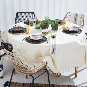 May include: A round, white tablecloth with fringed edges covers a wooden table with a black and white striped throw draped over a black wicker chair. The table is set with black and gold patterned plates, black placemats, and gold and green glasses. A small potted succulent plant sits in the center of the table.