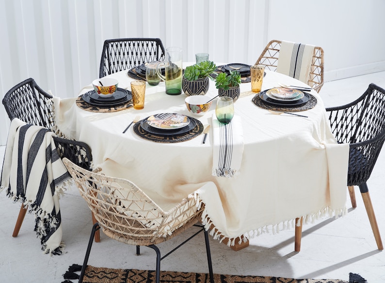 May include: A round, white tablecloth with fringed edges covers a wooden table with a black and white striped throw draped over a black wicker chair. The table is set with black and gold patterned plates, black placemats, and gold and green glasses. A small potted succulent plant sits in the center of the table.