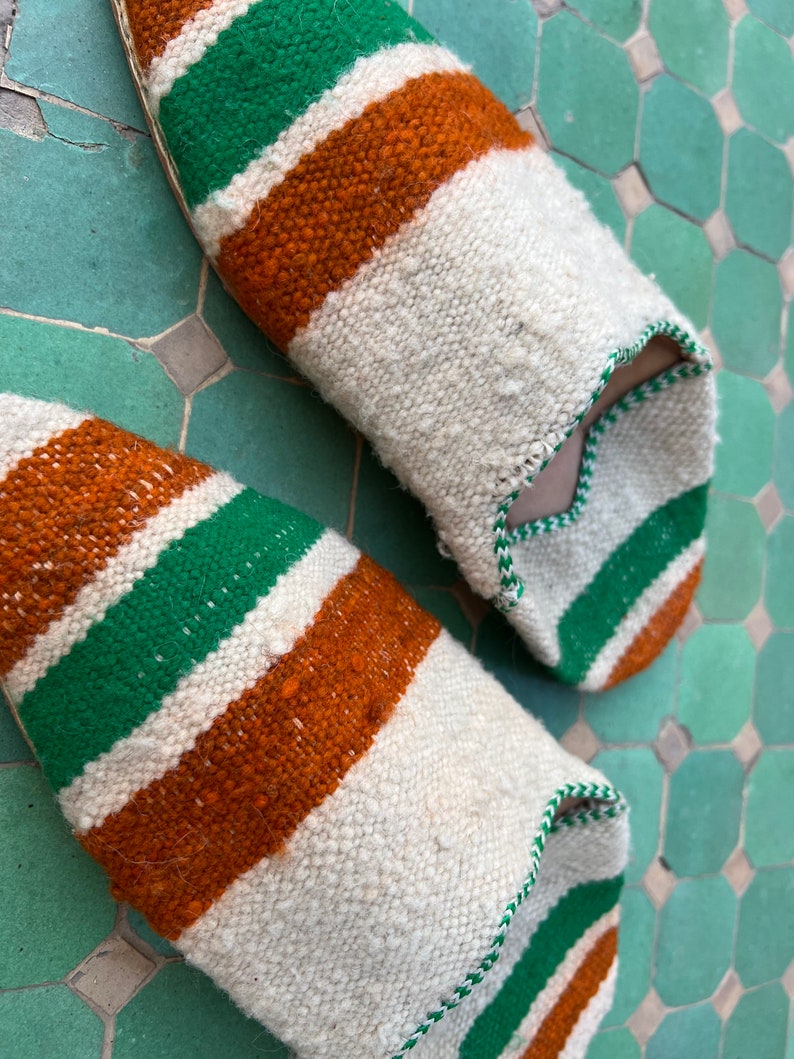 May include: A pair of white, green, and orange striped slippers. The slippers are made of a woven fabric and have a pointed toe.