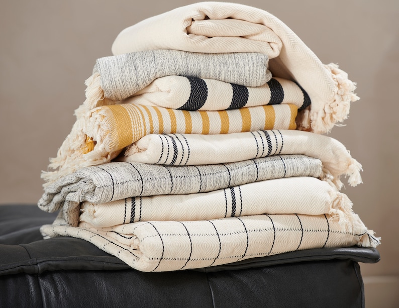 May include: A stack of eight woven throw blankets in various patterns and colors, including white, gray, black, and yellow. The blankets are folded and stacked on a black leather surface.