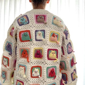 May include: A white crocheted cardigan with a colorful cat design. The cardigan is made up of squares with different colored borders and each square features a crocheted cat in a different color.