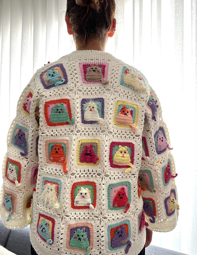 May include: A white crocheted cardigan with a colorful cat design. The cardigan is made up of squares with different colored borders and each square features a crocheted cat in a different color.