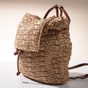 Può includere: Uno zaino in paglia intrecciata con cinturini e dettagli in pelle marrone. La borsa ha una chiusura con patta e una parte superiore con coulisse. Lo zaino è di colore marrone e marrone e sembra essere realizzato con materiali naturali.