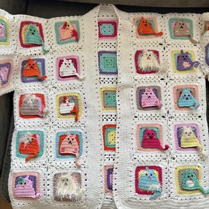 May include: A white crocheted cardigan with a variety of colorful crocheted cats attached to the front. The cardigan has a long sleeve and an open front.