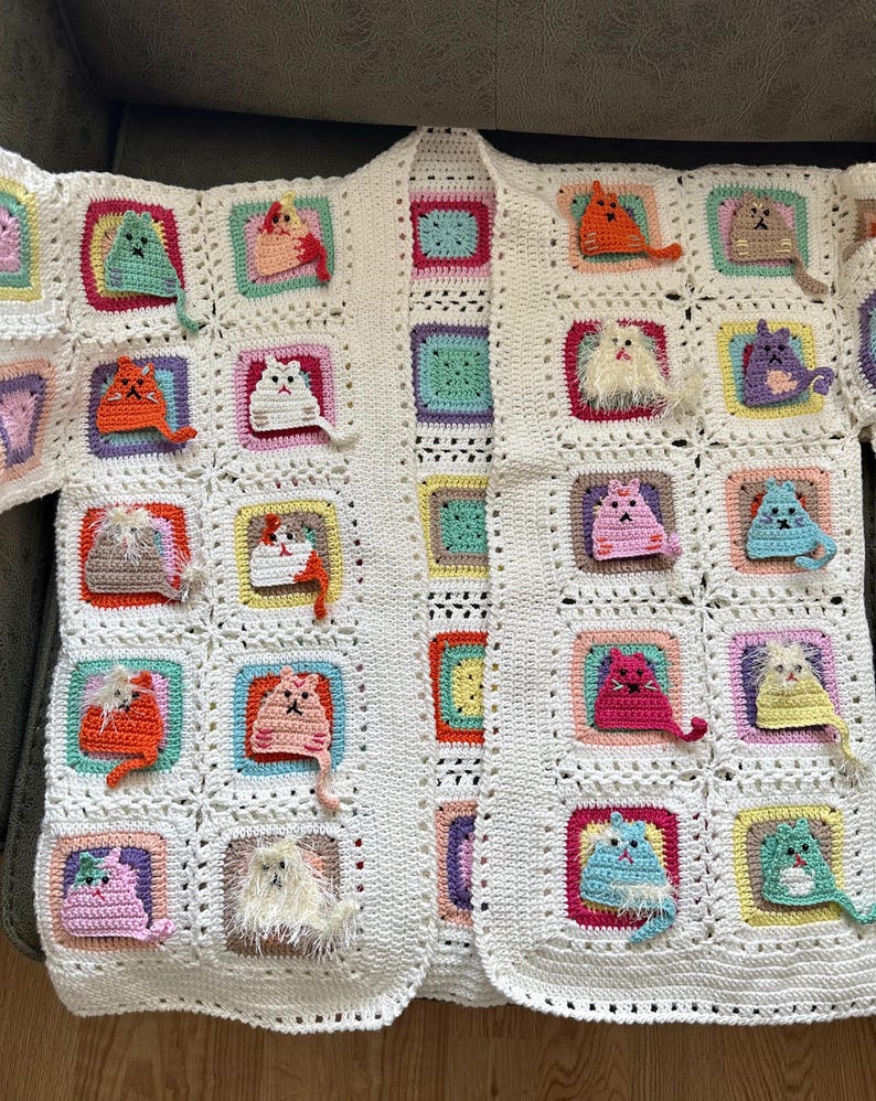 May include: A white crocheted cardigan with a variety of colorful crocheted cats attached to the front. The cardigan has a long sleeve and an open front.