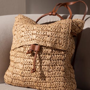 Puede incluir: Una mochila de paja tejida con correas de cuero marrón y cierre de cordón. La bolsa tiene un cierre de solapa y una apariencia natural texturizada. La mochila está diseñada para transportar objetos.