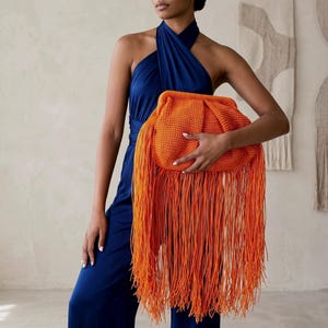 Puede incluir: Un bolso de mano naranja con largas borlas se sostiene contra un mono azul oscuro. El bolso tiene una textura tejida y una forma redondeada. Las largas borlas cuelgan, creando un contraste visual audaz. El fondo es de color neutro.