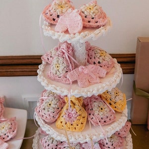 May include: A three-tiered white cake stand with crocheted baby shower favors. The favors are in the shape of small bags and dresses, with pink, yellow, and white colors.