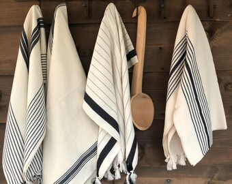 May include: Four white kitchen towels with black stripes hanging on hooks. The towels have fringe at the bottom and are made of a soft, absorbent material.