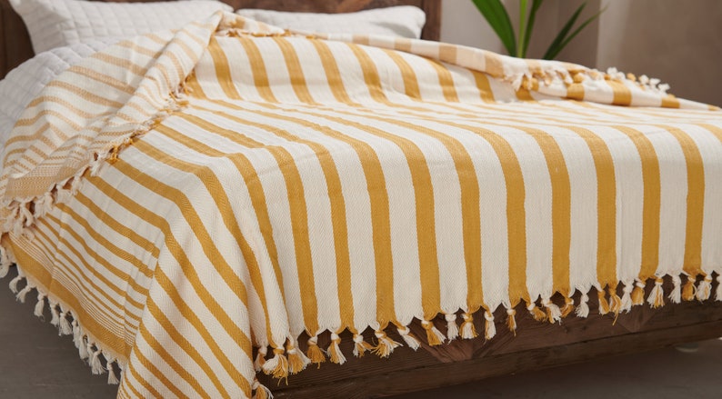 May include: A yellow and white striped throw blanket with tassels on the edges. The blanket is folded on a wooden bed frame.