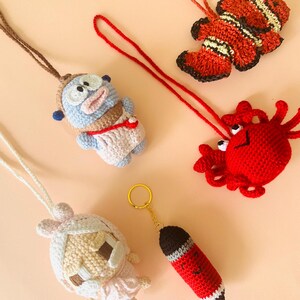 May include: A selection of handmade crochet items, featuring a blue cartoon character, a clownfish, a red crab, a white doll, and a red pencil keychain. Each item is made with detailed stitching and bright colours, ideal for gifts or collecting.