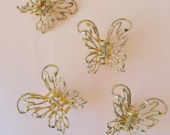 May include: Four gold-colored butterfly hair clips. Each clip features a delicate, openwork design with a spring mechanism. The clips are arranged on a light pink background, showcasing their intricate details and metallic sheen.