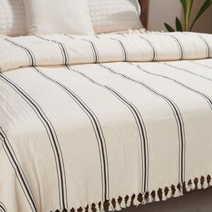 May include: A white and black striped throw blanket with fringe on the edges. The blanket is folded on a bed with white pillows.