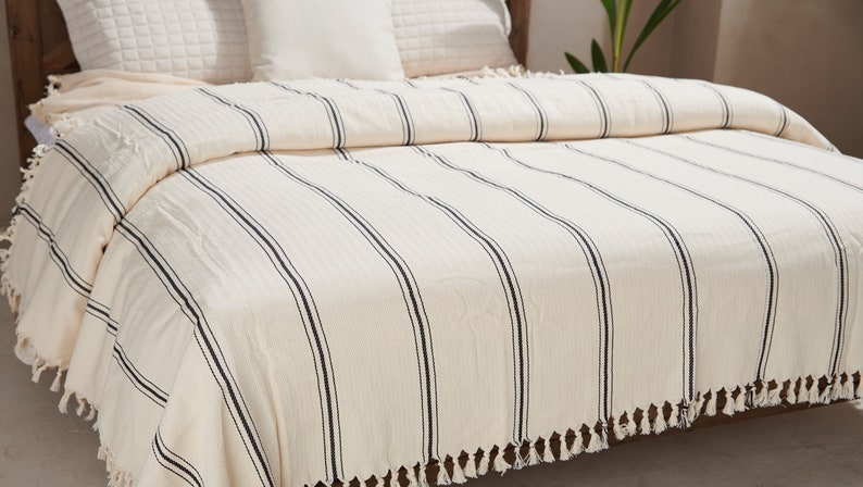 May include: A white and black striped throw blanket with fringe on the edges. The blanket is folded on a bed with white pillows.