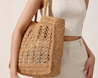 May include: A tan crochet tote bag with a rectangular shape and two handles. The bag has an open weave design, revealing the contents inside. The bag is carried by a person wearing a cream-coloured outfit.