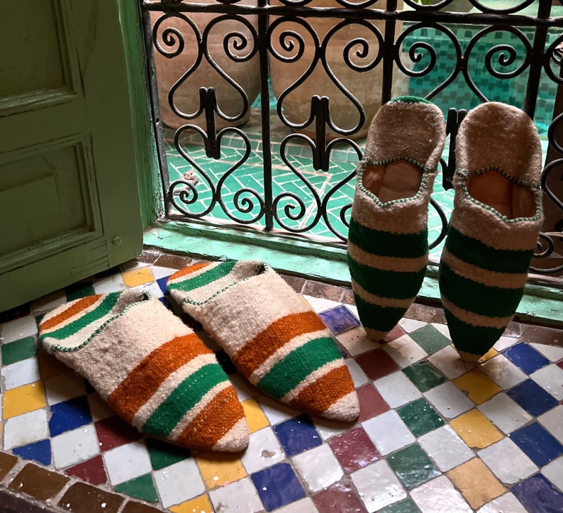 May include: Four pairs of striped slippers in green, white, and orange. The slippers are made of a soft, woven fabric and have a pointed toe.