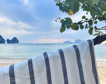 May include: A striped beach towel with alternating navy blue and white vertical stripes hangs from a tree branch. The towel is on a sandy beach with the ocean and sky in the background. The towel has thin red and blue accent stripes.