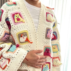 May include: A white crocheted cardigan with a colorful cat design. The cardigan has a V-neck and long sleeves. The cat design is made up of different colored squares with a cat in each square.