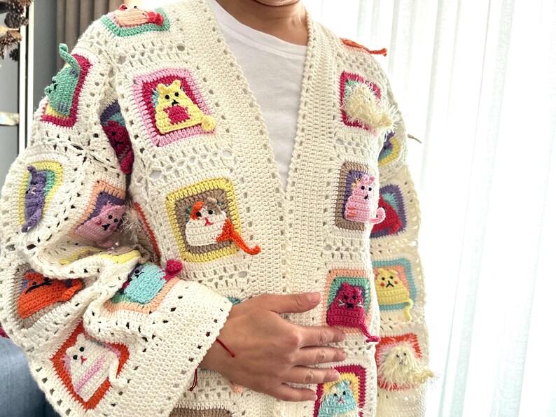 May include: A white crocheted cardigan with a colorful cat design. The cardigan has a V-neck and long sleeves. The cat design is made up of different colored squares with a cat in each square.