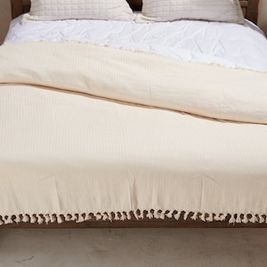 May include: Cream-colored bedspread with a textured, ribbed design and a fringe of tassels along the bottom edge. The bed is made up with white bedding and pillows. The bed frame is made of dark wood.