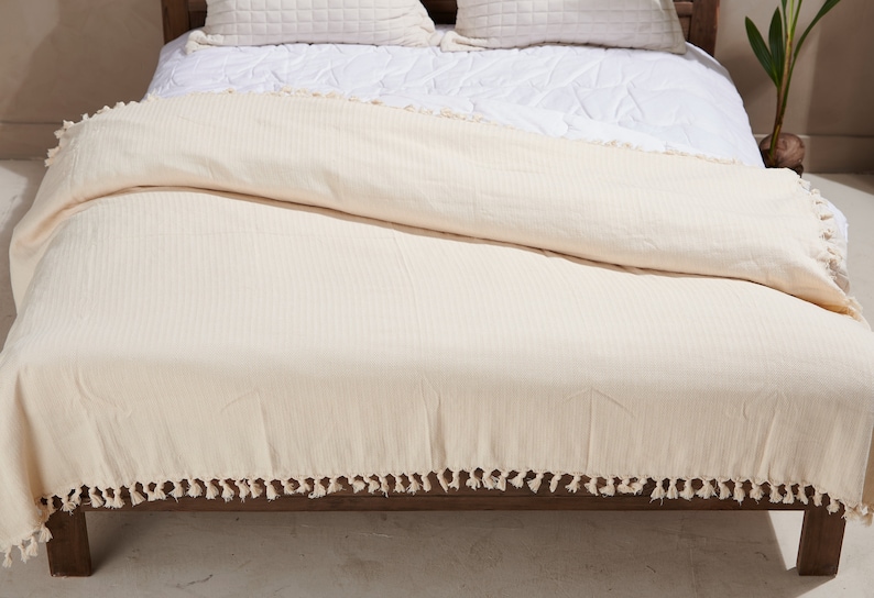 May include: A cream-colored waffle weave throw blanket with a fringe border. The blanket is folded on a bed with white bedding.