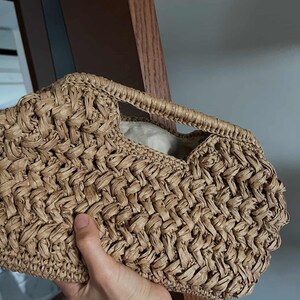 May include: A woven handbag in a light brown hue. The bag features a textured, basket-weave design and a small handle. The interior is lined with a light-colored fabric. The bag is held by a person.