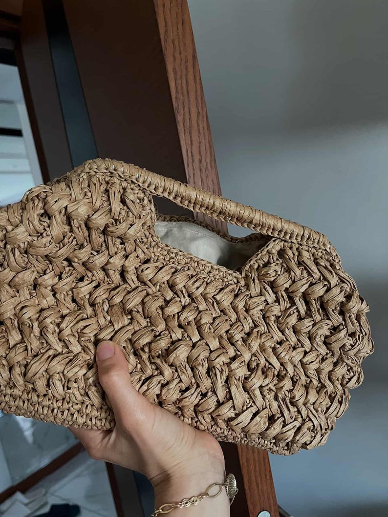 May include: A woven handbag in a light brown hue. The bag features a textured, basket-weave design and a small handle. The interior is lined with a light-colored fabric. The bag is held by a person.