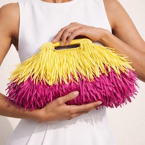 May include: A vibrant, fringed handbag in yellow and magenta. The top handle and upper section are yellow, transitioning to magenta fringe below. The bag is held against a white background.