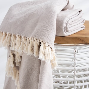 Peut inclure: Une couverture en laine gris clair avec un motif en chevrons et une bordure en franges. La couverture est pliée et drapée sur une table en osier blanche.