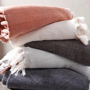 Può includere: Una pila di cinque coperte a spina di pesce in vari colori: corallo, bianco, marrone scuro, grigio chiaro e blu navy. Ogni coperta ha bordi con frange.