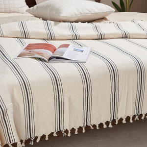 May include: A white throw blanket with black stripes and tassels. The blanket is on a bed with pillows and a magazine on top.
