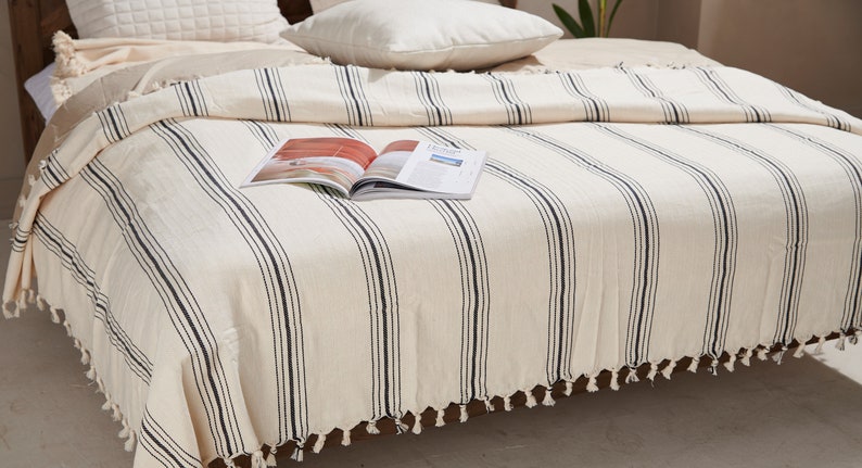 May include: A white throw blanket with black stripes and tassels. The blanket is on a bed with pillows and a magazine on top.