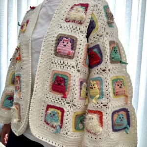 May include: A white crocheted cardigan with colorful cat appliques. The cardigan has a square pattern and features cats in various colors, including pink, red, yellow, and blue.