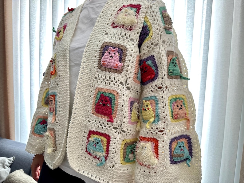 May include: A white crocheted cardigan with colorful cat appliques. The cardigan has a square pattern and features cats in various colors, including pink, red, yellow, and blue.