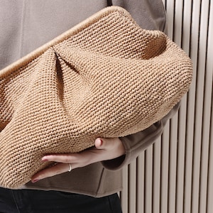 Könnte beinhalten: Eine gewebte Clutch-Tasche in hellbrauner Farbe mit leicht strukturierter Oberfläche. Die Tasche ist leicht aufgebläht und hat eine abgerundete Form.