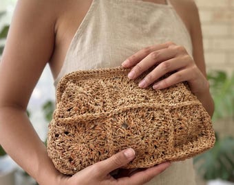 Puede incluir: Un bolso de mano rectangular confeccionado en fibra de rafia de color tostado natural, con una estructura rígida y un patrón de ganchillo de cuadrados repetitivos con motivos florales complejos en toda la superficie.