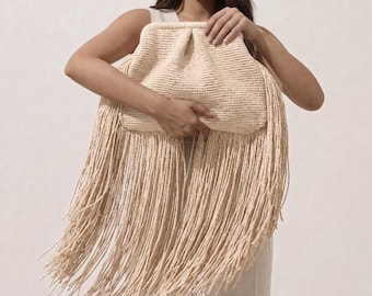May include: A beige woven clutch bag featuring a structured frame closure and a prominent, long fringe detail that extends from the base of the bag. The texture of the material is consistent throughout the body and the hanging fringe strands.