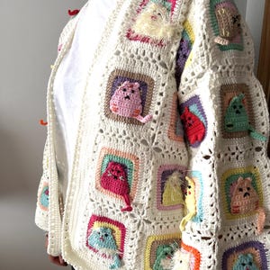 May include: A white crocheted cardigan with colorful cat appliques. The cardigan has a V-neck and long sleeves. The cat appliques are in various colors, including pink, red, yellow, green, and blue.