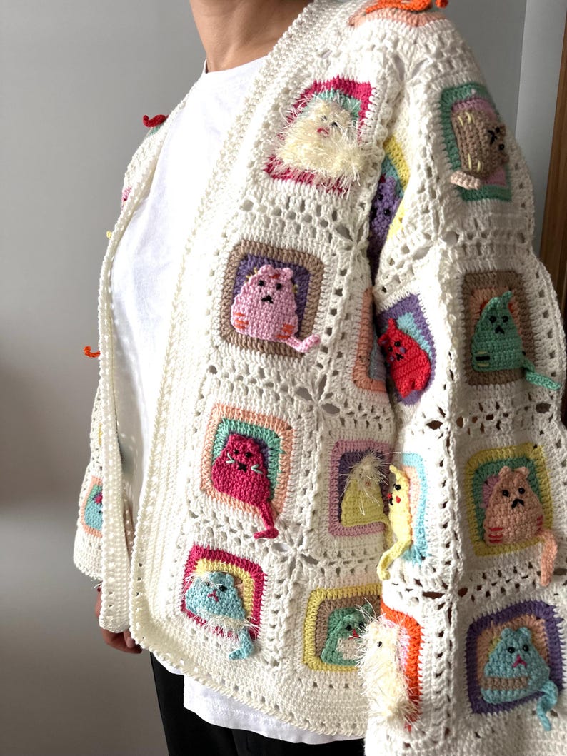 May include: A white crocheted cardigan with colorful cat appliques. The cardigan has a V-neck and long sleeves. The cat appliques are in various colors, including pink, red, yellow, green, and blue.