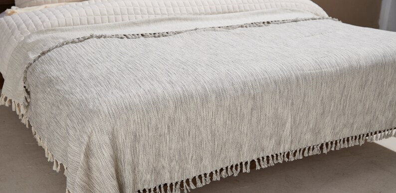 May include: A white throw blanket with a subtle striped pattern and fringe trim. The blanket is draped over a bed.
