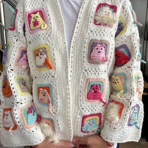 May include: White crochet cardigan with colorful cat appliques. The cardigan has a loose fit and a long sleeve. The cat appliques are crocheted in different colors and have a 3D effect.