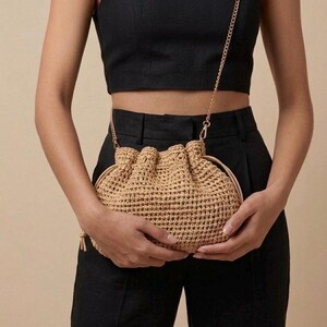 May include: A beige, woven handbag with a drawstring closure and a gold chain strap. The bag is held by a person wearing a black crop top and black trousers. The bag's texture is visible.
