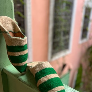 May include: A pair of green and white striped slippers with pointed toes. The slippers are made of a soft, woven fabric and are resting on a wooden surface.